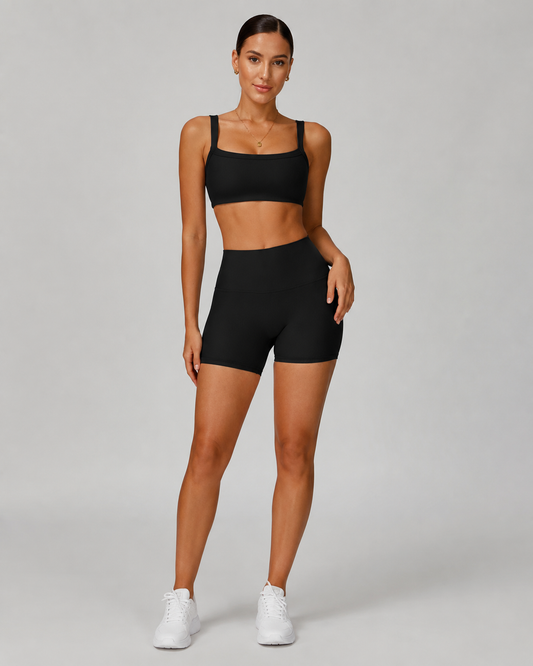 A woman modeling a black activewear set consisting of a square-neck sports bra and high-waisted biker shorts, paired with white sneakers, against a solid light gray studio background.