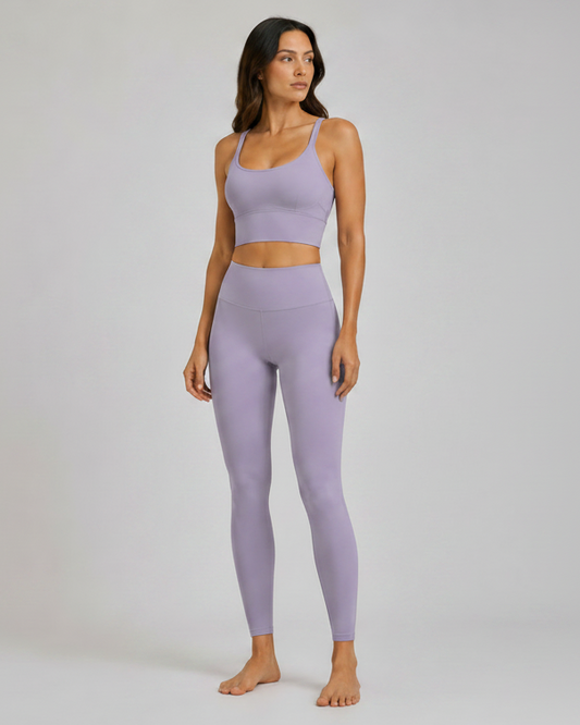 A woman modeling a matching lavender purple activewear set consisting of a sports bra and high-waisted leggings, standing against a neutral light gray studio background.