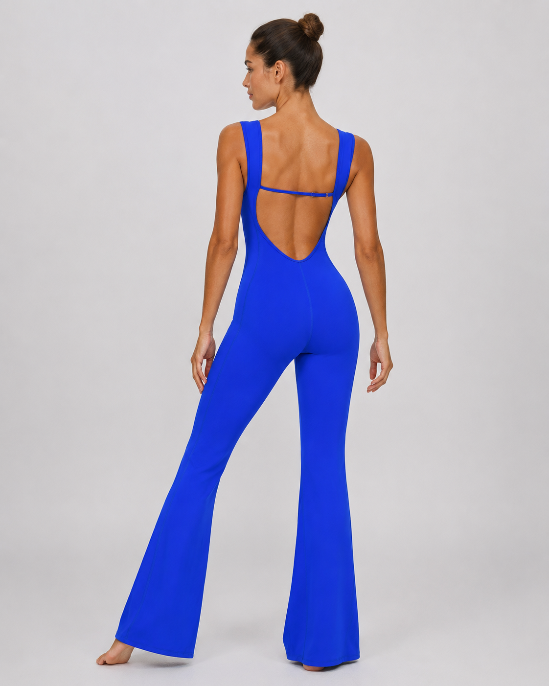 A woman modeling a bright royal blue one-piece jumpsuit from the back, featuring a low open back with thin straps and wide flared legs, set against a plain light gray studio background.