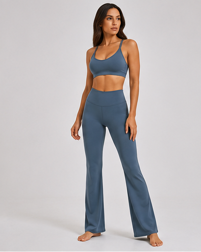 A woman modeling a matching two-piece activewear set consisting of a slate blue sports bra and high-waisted flare leggings, photographed against a neutral light gray studio background.