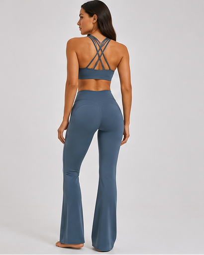 A woman shown from the back wearing a matching slate blue activewear set consisting of a sports bra with crisscross straps and high-waisted flared leggings, set against a plain light gray studio background.